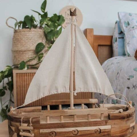 Wooden model sailboat with white sail on a wooden surface, surrounded by plants and decor.