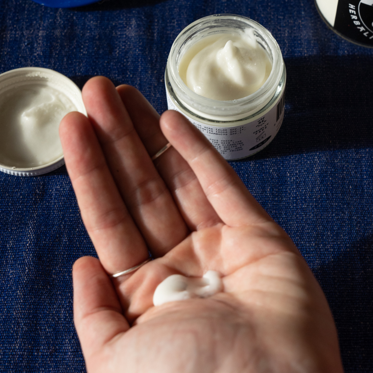 Hand holding a small amount of white cream with jars of cream in the background on a blue textured surface.