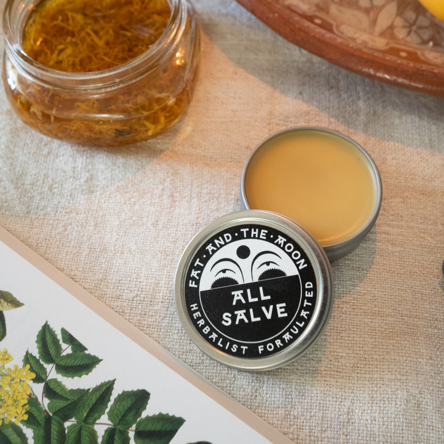 All Salve container with lid open, showing a herbalist-formulated balm, on a textured surface with a jar of oil and botanical illustration.