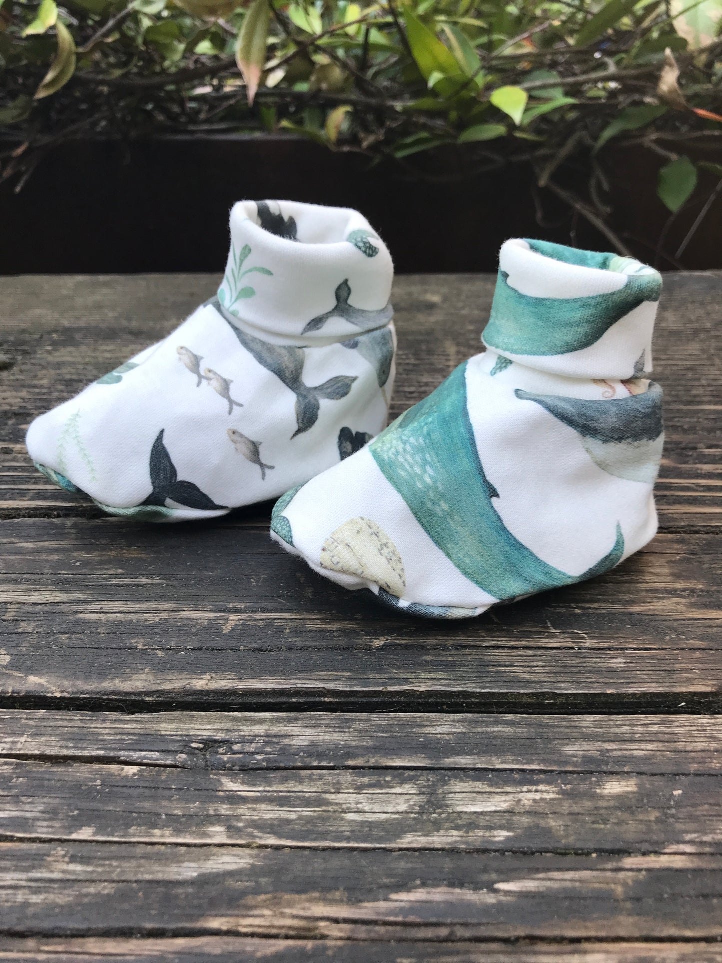 infant baby whale print booties that are white with blue wales printed on them sitting on a wood bench with some leaves in the background