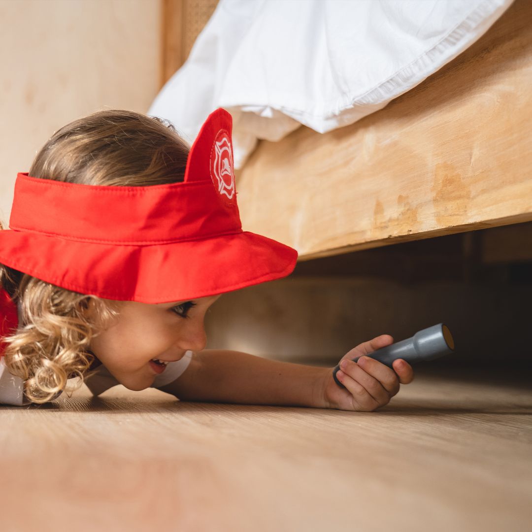Child wearing a red visor and holding a flashlight, looking under a bed.