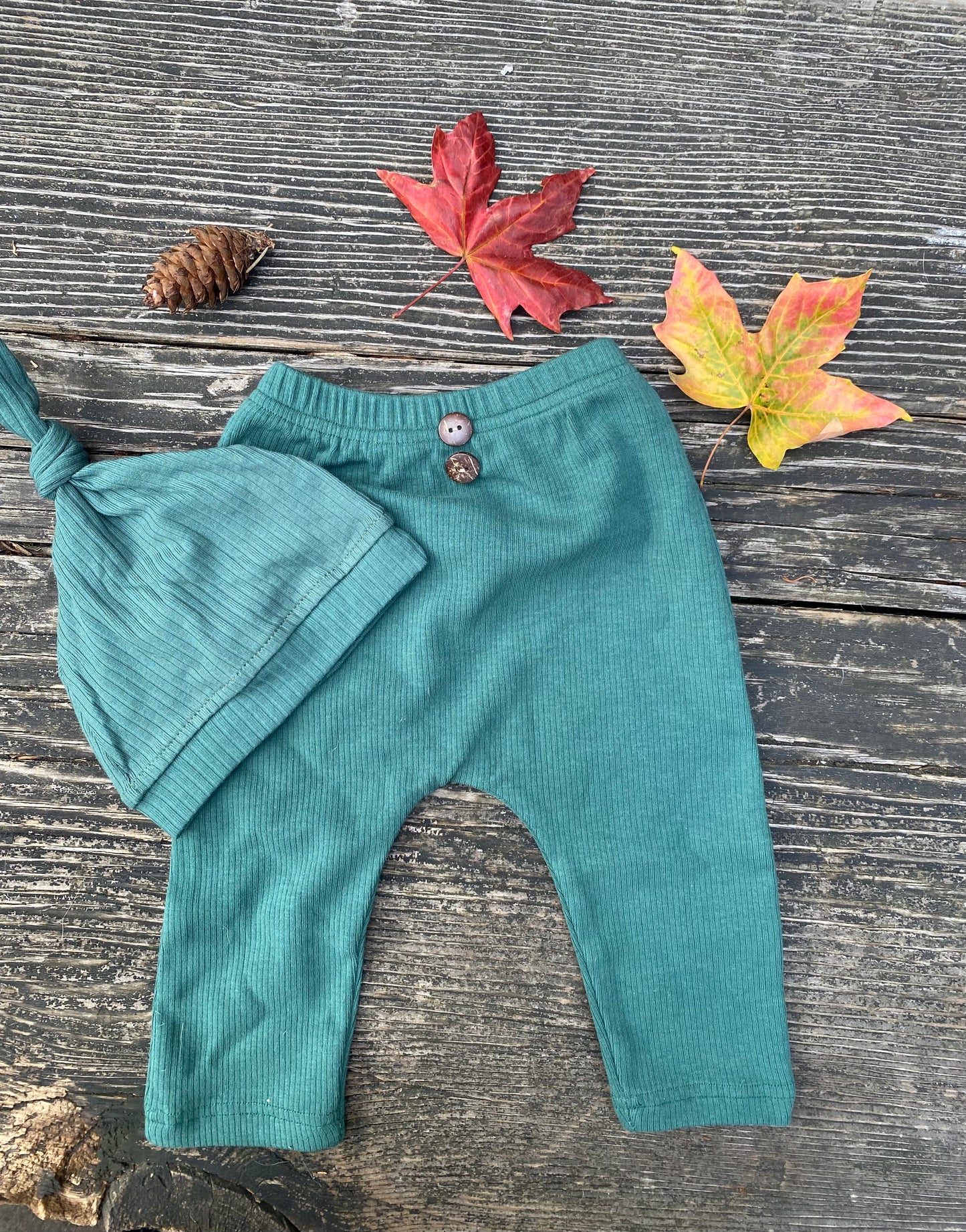 Green baby pants and hat on a wooden surface with autumn leaves and a pine cone.