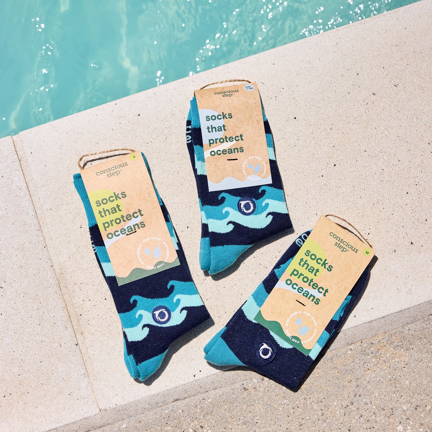 Three pairs of socks with ocean-themed design and 'socks that protect oceans' packaging by Oceana on a concrete surface near water.