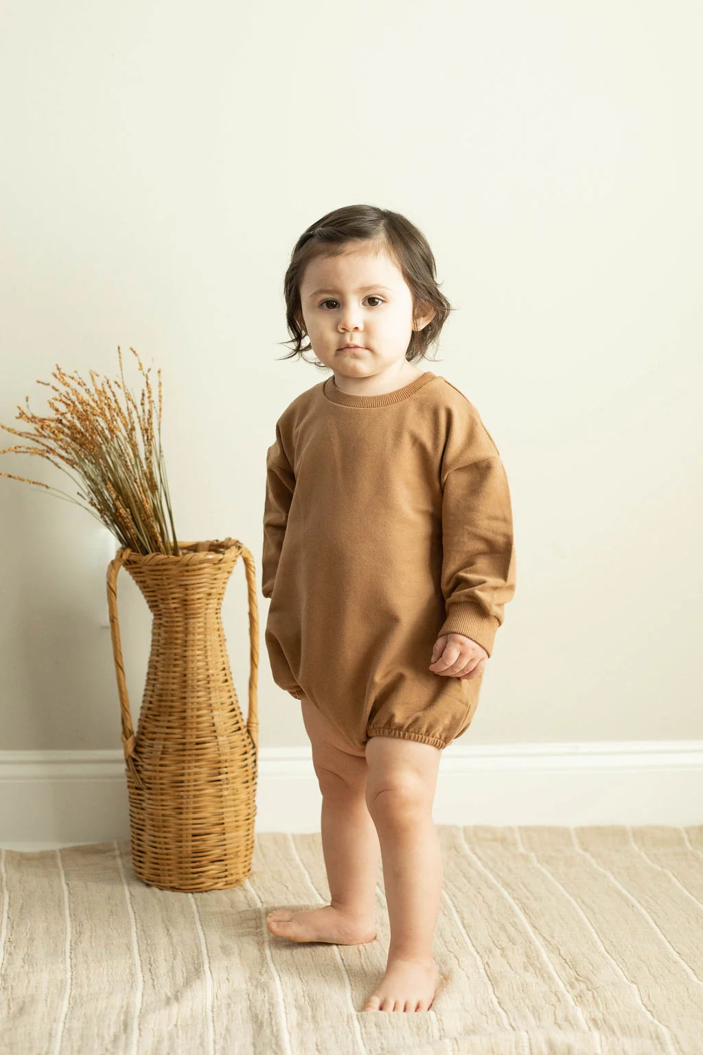 Child wearing a brown outfit standing next to a woven basket indoors.