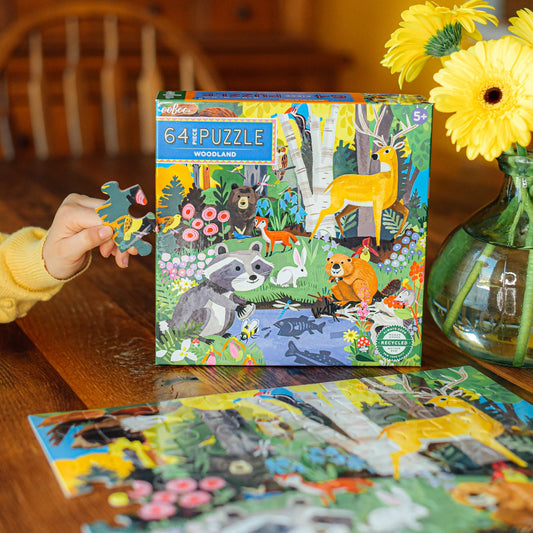 child wearing a yellow sweater holding a puzzle piece next to the box for Woodland 64-piece Puzzle