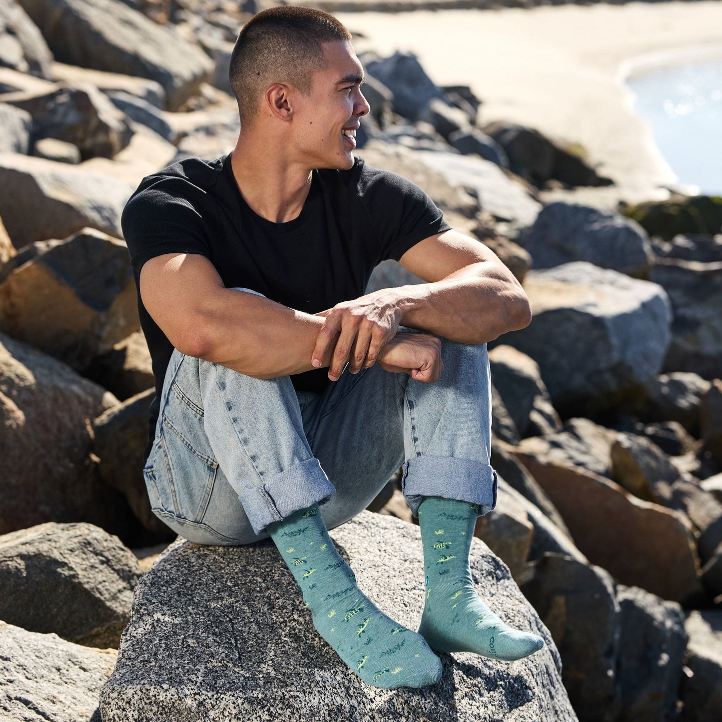 Man sitting on rocks wearing black t-shirt, light blue jeans, and teal socks with yellow patterns.