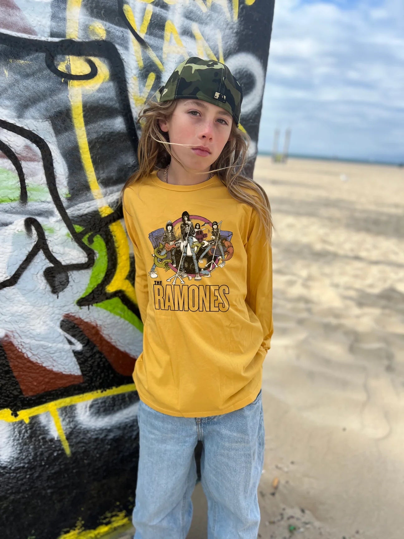 Child standing in front of a graffiti wall wearing jeans, a camouflage bandana, and a Ramones organic long sleeve tee