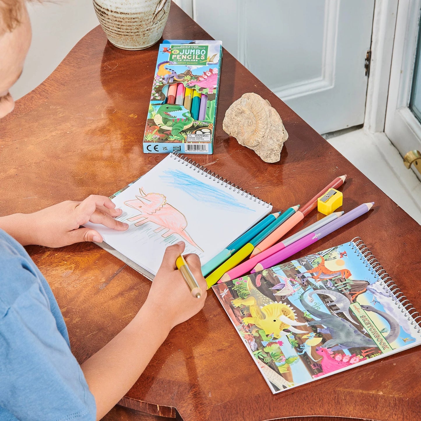 child at a table drawing on a dinosaur square sketchbook