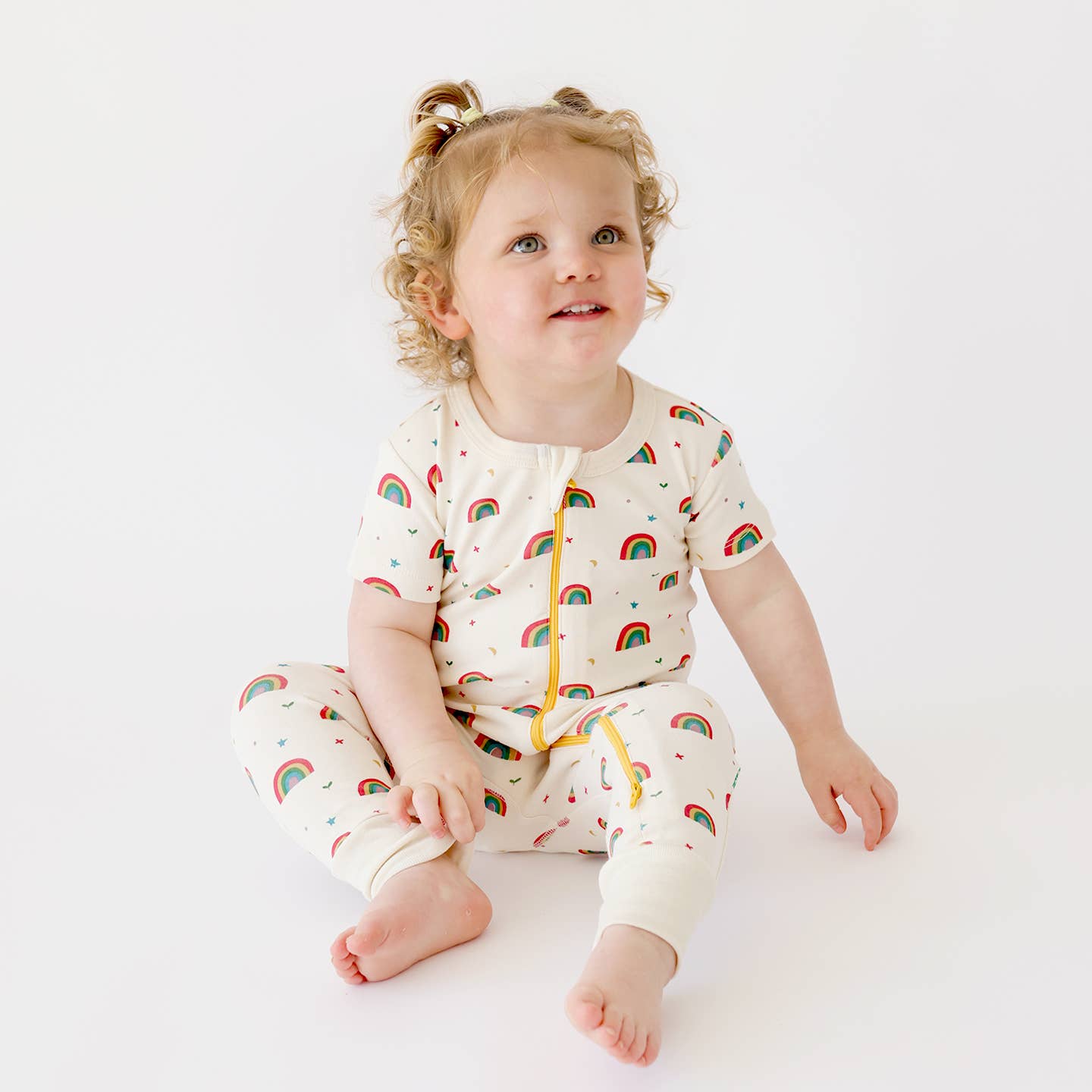 Toddler with curly blonde hair with two pig tales sitting and looking up while wearing parade organics Short Sleeve Rainbow Romper w/ 2-way Zipper