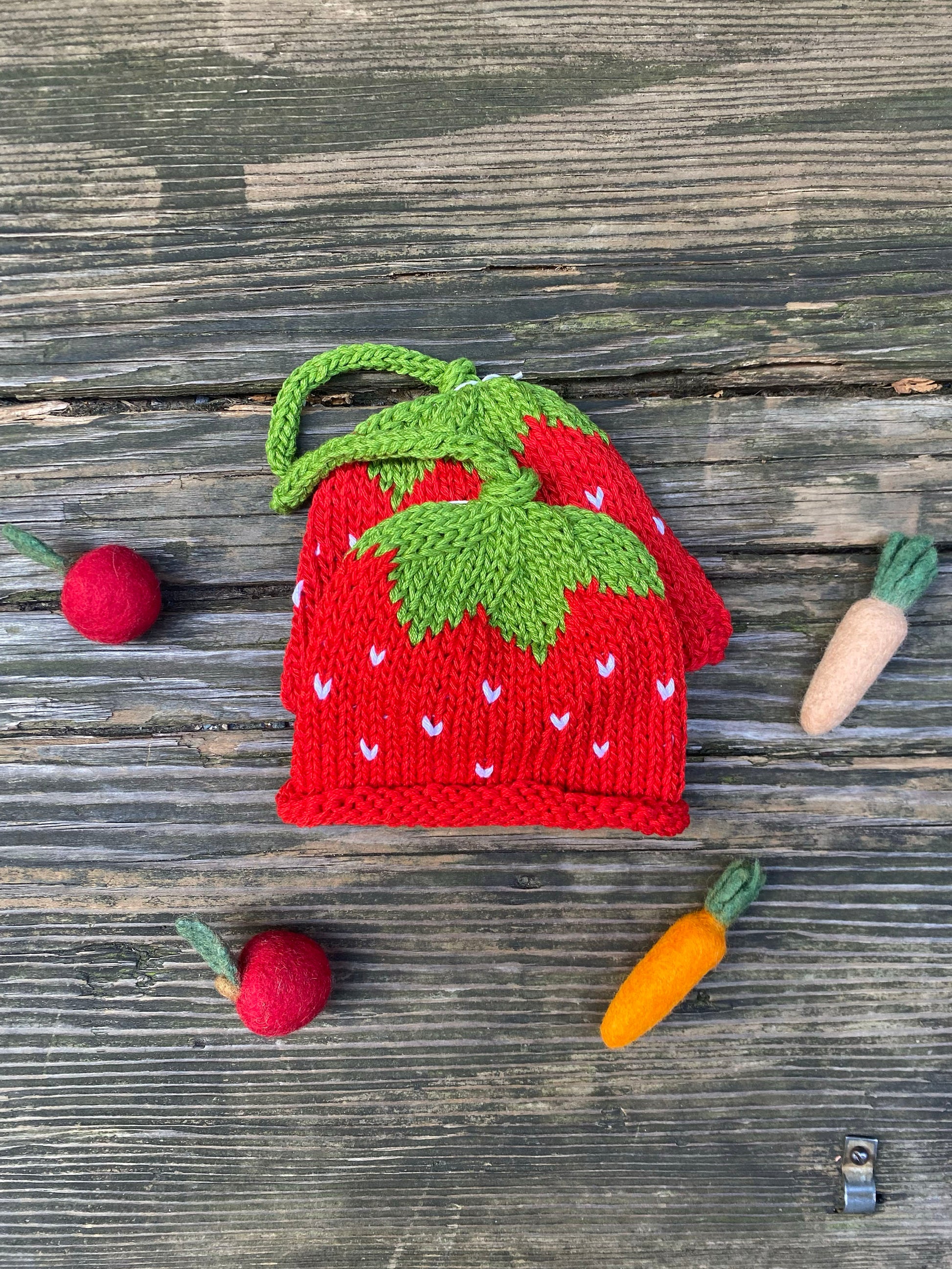 Red strawberry-shaped knitted hat with green leaves on a wooden surface.