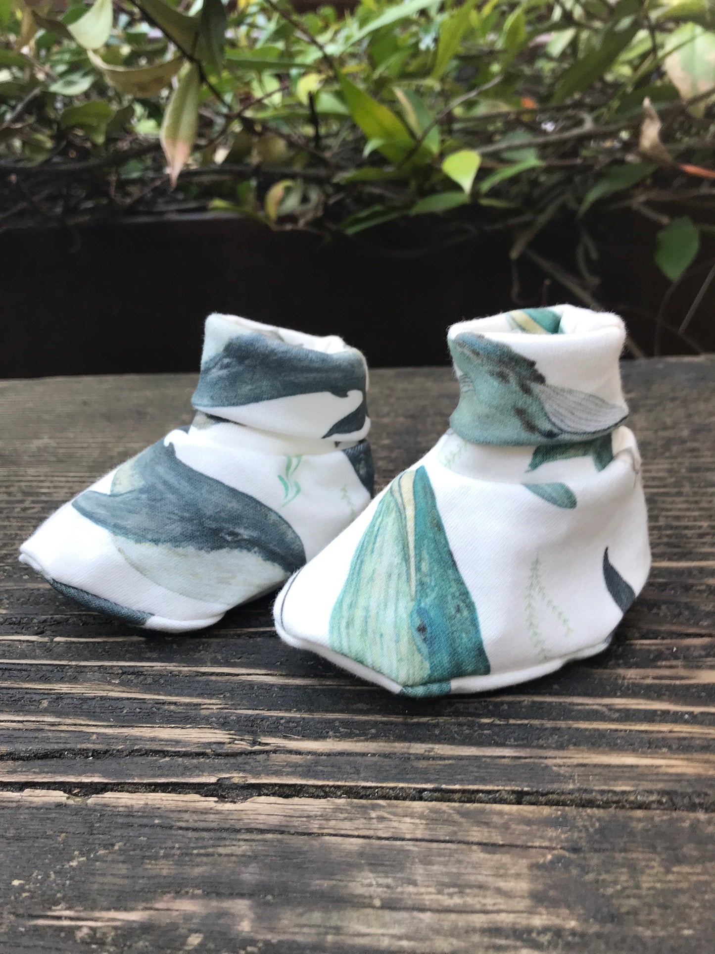 infant baby booties that are white with blue wales printed on them sitting on a wood bench with some leaves in the background