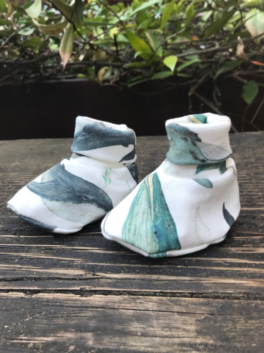 infant baby booties that are white with blue wales printed on them sitting on a wood bench with some leaves in the background