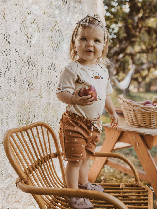 child standing on a chair outside holding an apple wearing terracotta moon organic shorts