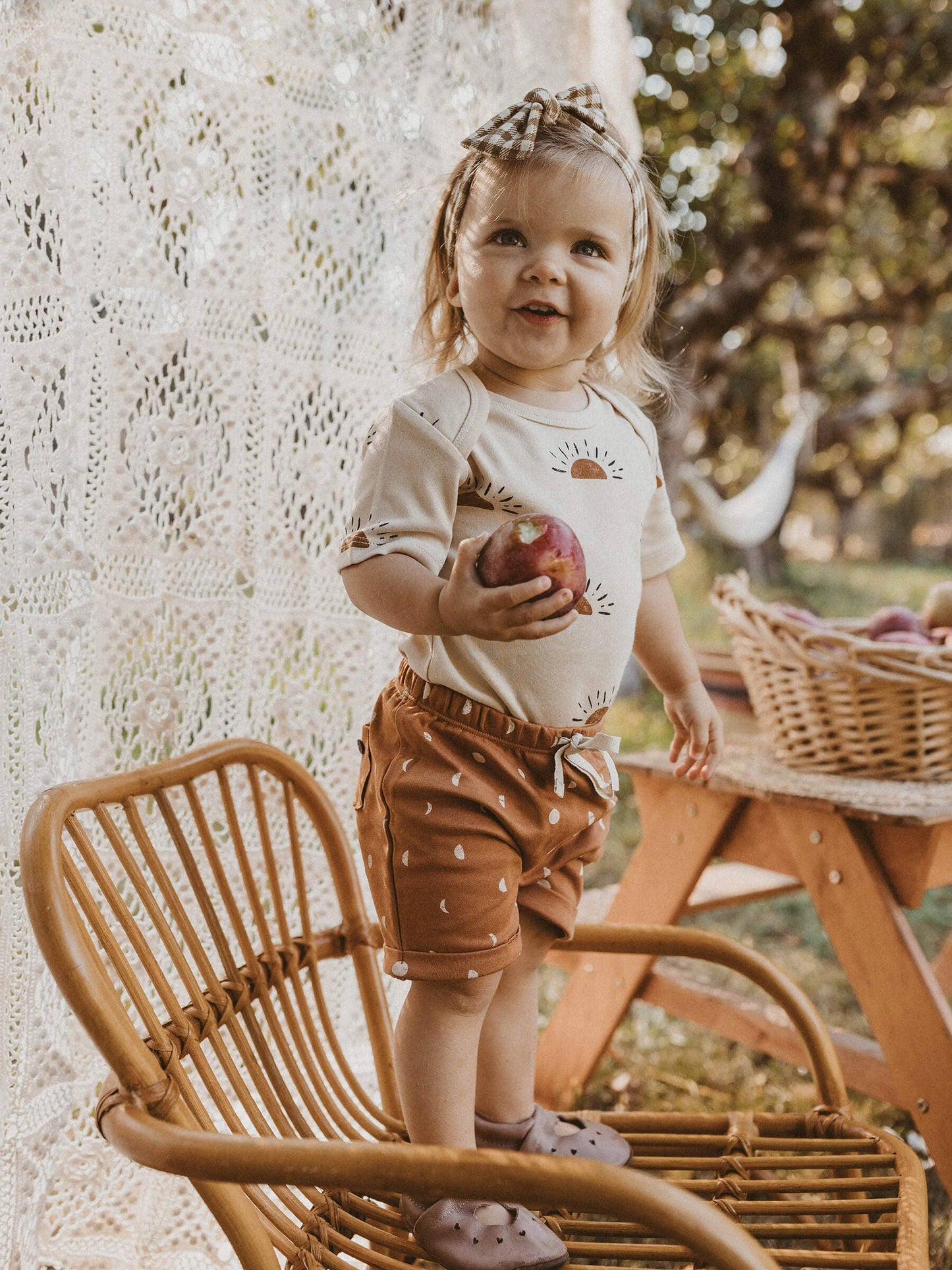 child standing on a wooden chair holding a pomegranate waring sun organic short sleeve onesie