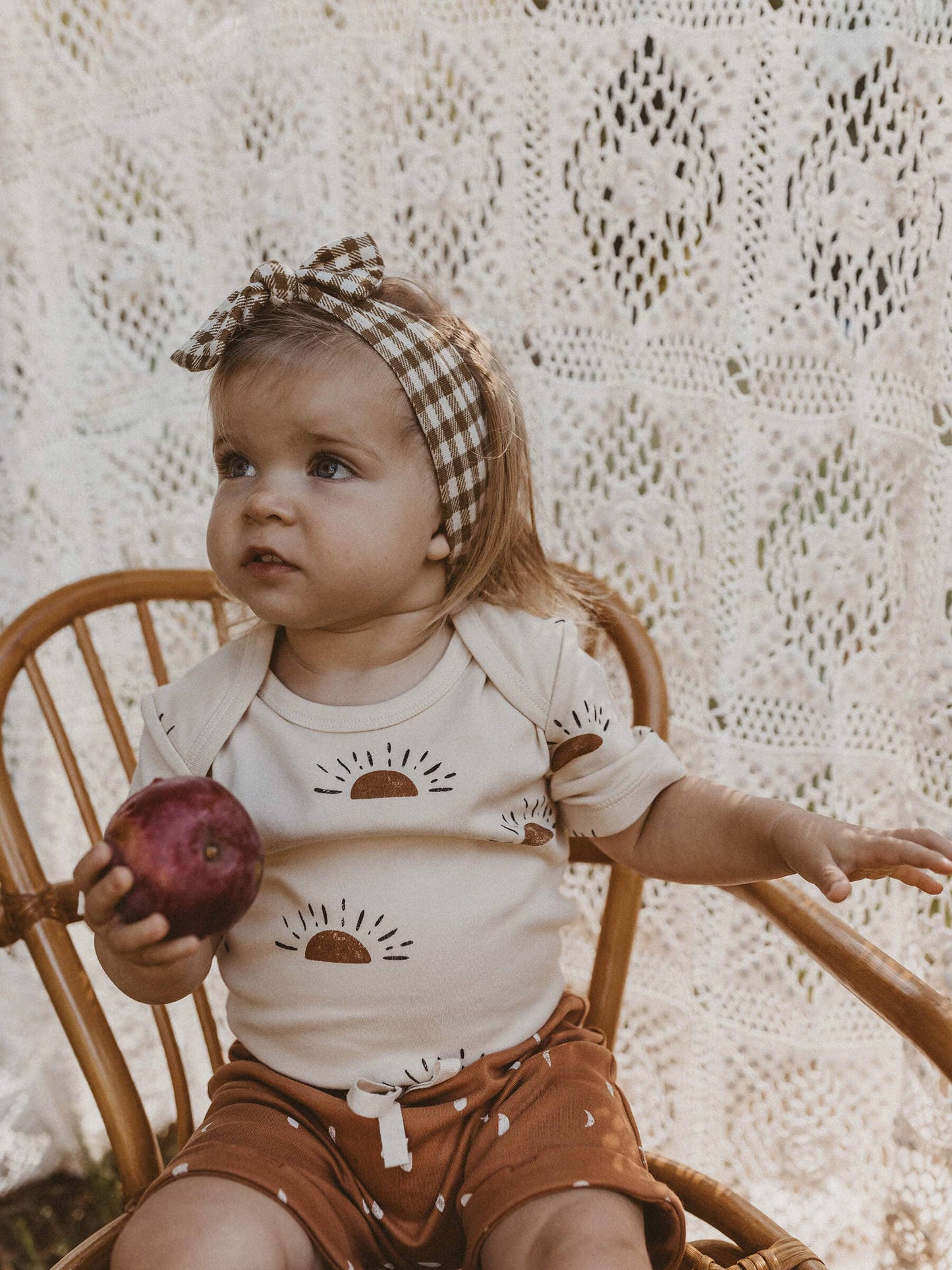 child sitting in a chair holding a pomegranate wearing sun organic short sleeve onesie