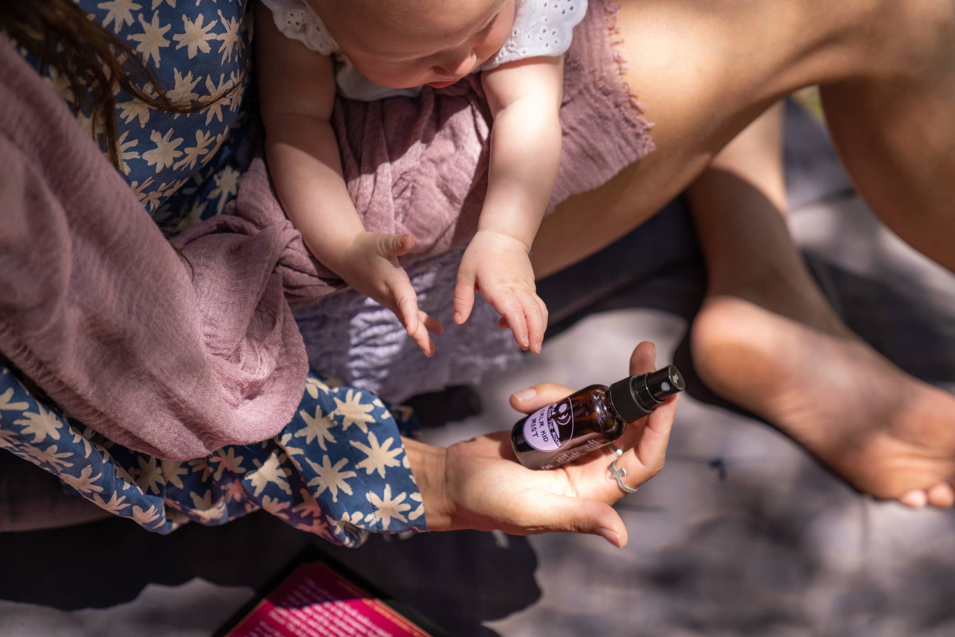 a person sitting on a blanket outdoors, holding a baby and a kid calm mist bottle in their hand