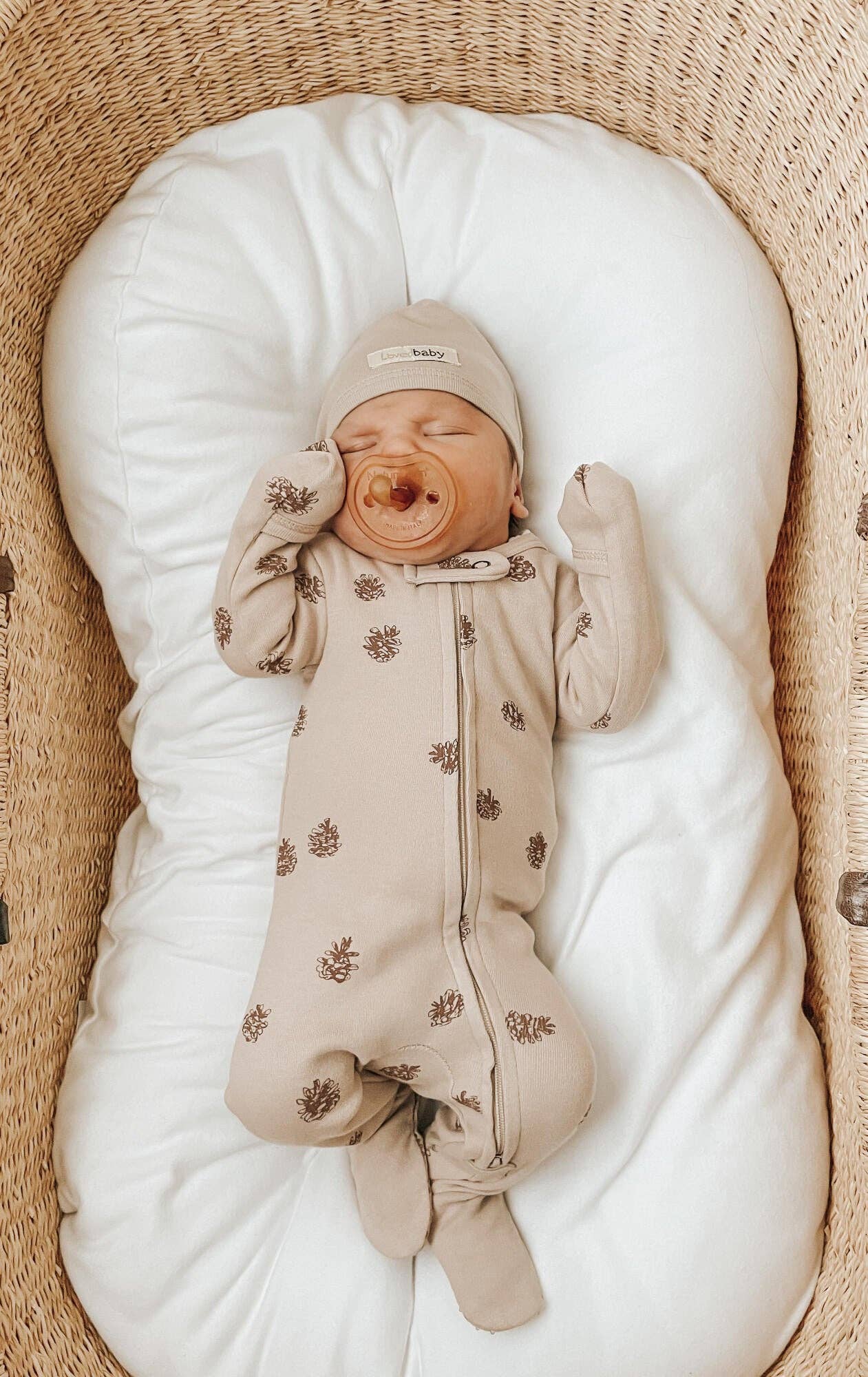 Baby in a beige footie with pinecones patterns lying on a white cushion in a wicker crib.