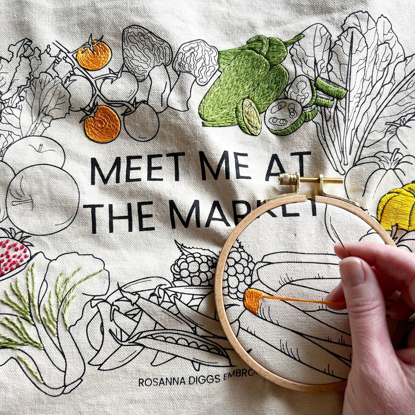 Embroidery hoop with embroidered vegetables and text 'Meet Me at the Market' on fabric.