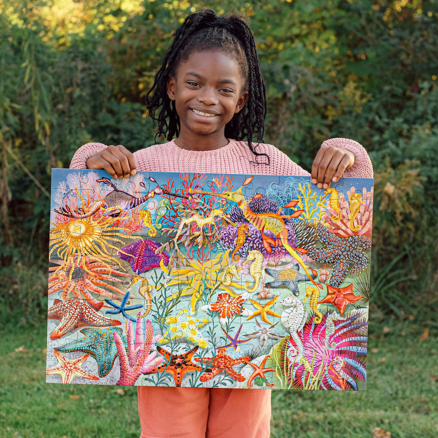 a child standing outside holding a completed seahorse, sea stars & sea slugs 100-piece puzzle
