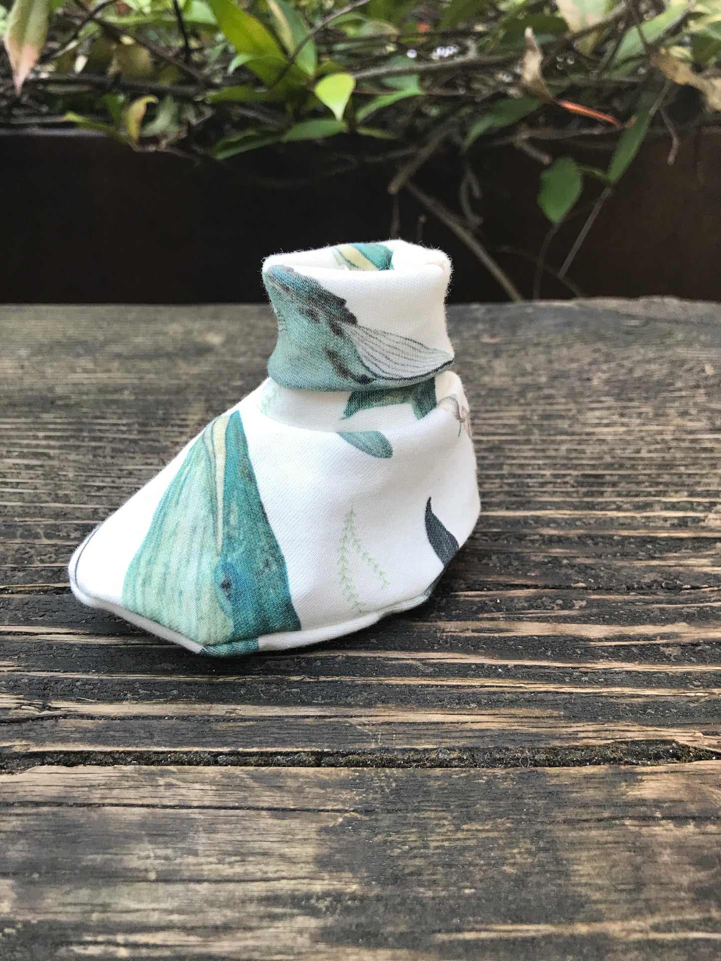 infant baby bootie that is white with blue wales printed on them sitting on a wood bench with some leaves in the background