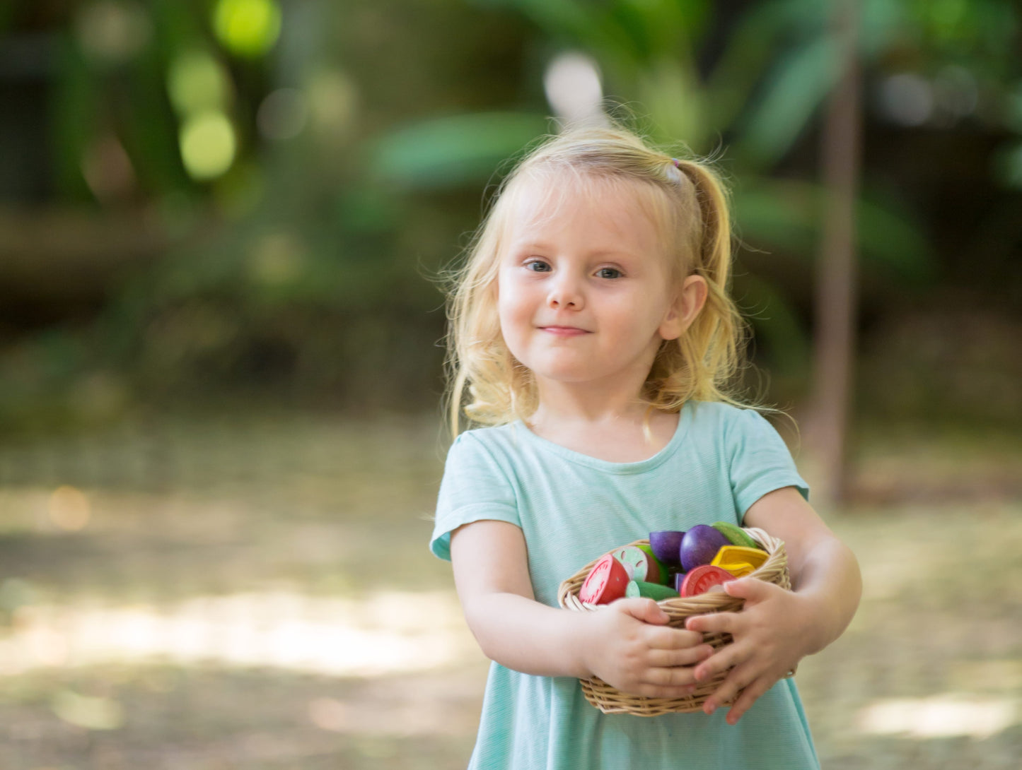 child standing out side holding a basket with a fruit and vegetable play set by plan toys