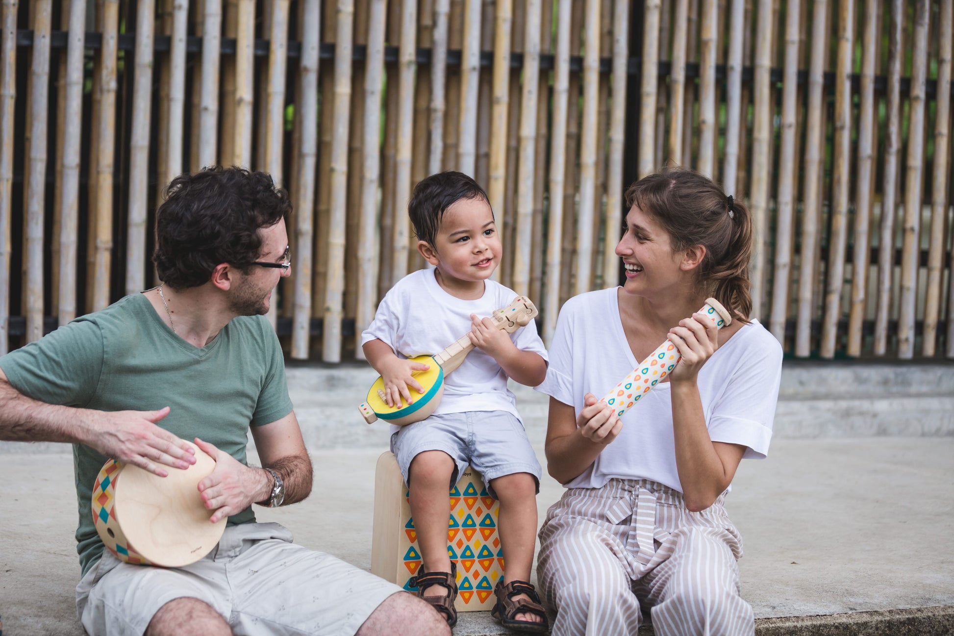 two adults and a child sitting outside playing with toy musical instruments by PlanToys