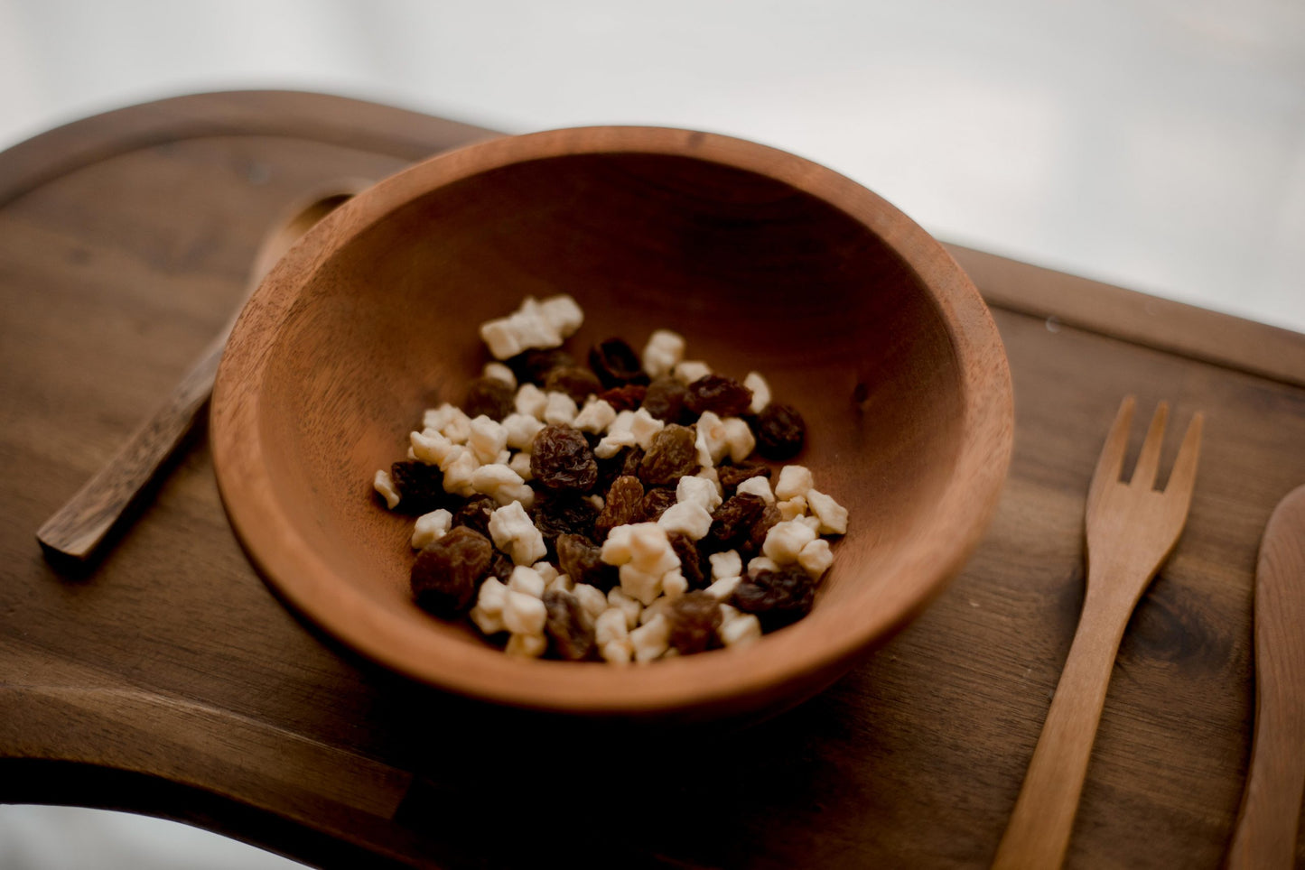 Wooden toddler bowl with utensils on a wooden tray. There is raisins in the bowl