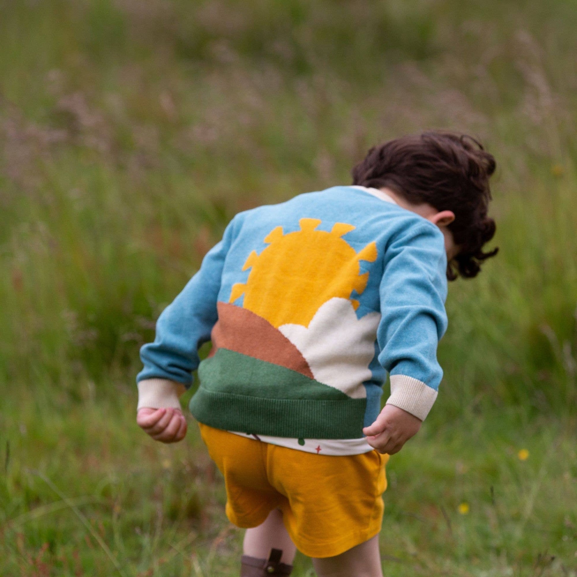 Child walking in a grass field wearing From One To Another Sunshine Design Knitted Cardigan