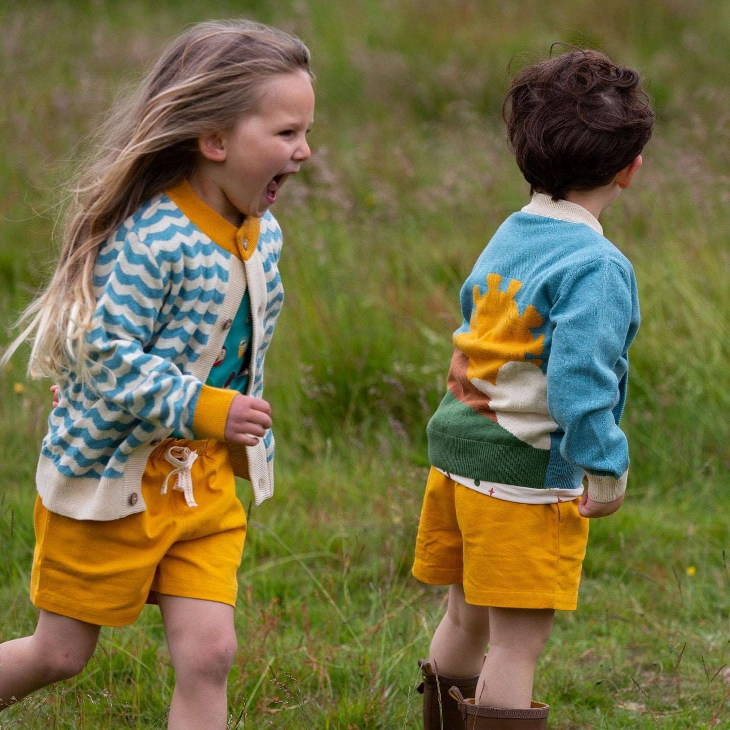 Two children walking in a grass field, one wearing From One To Another Sunshine Design Knitted Cardigan
