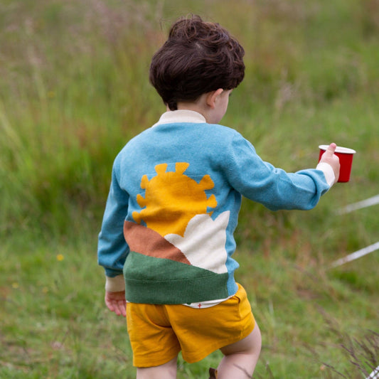 Child walking in a grass field holding a red cup wearing From One To Another Sunshine Design Knitted Cardigan