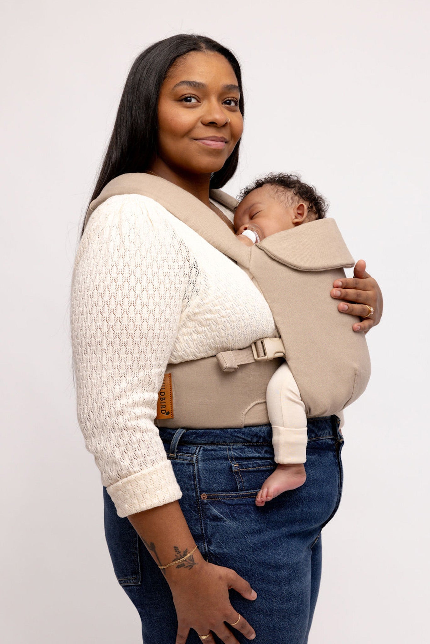 Woman holding a baby in a beige baby carrier against a plain background