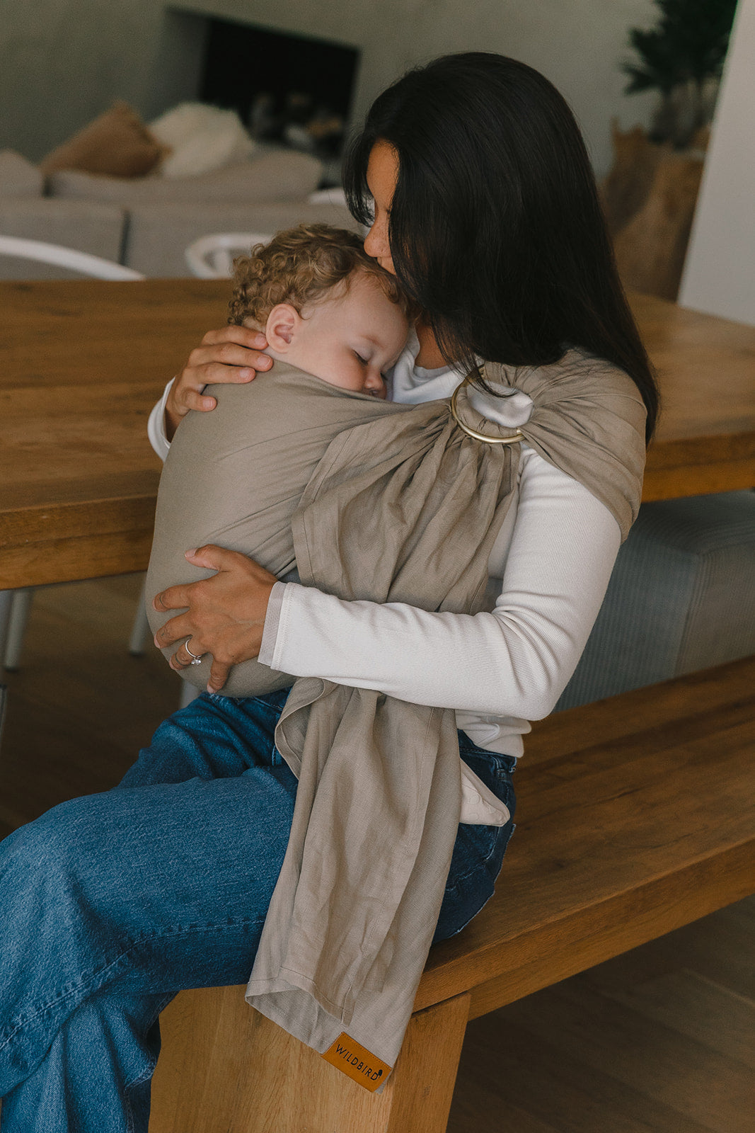 Woman holding a baby in a sling on a wooden bench indoors