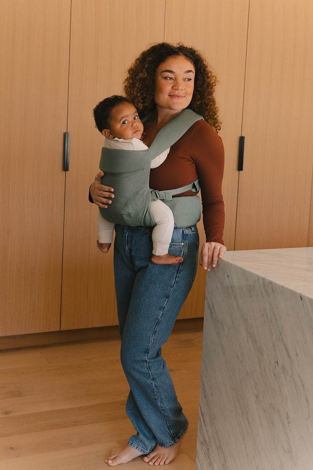 Woman holding a baby in a carrier in a room with wooden walls and a concrete floor.
