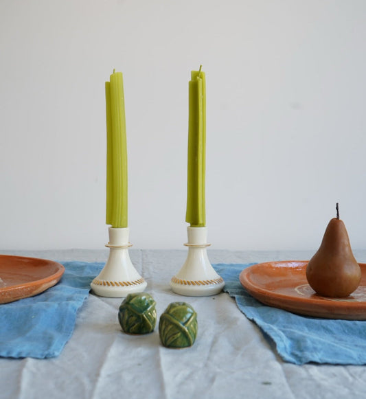Two green candles in white candlesticks on a table with a pear and decorative items.