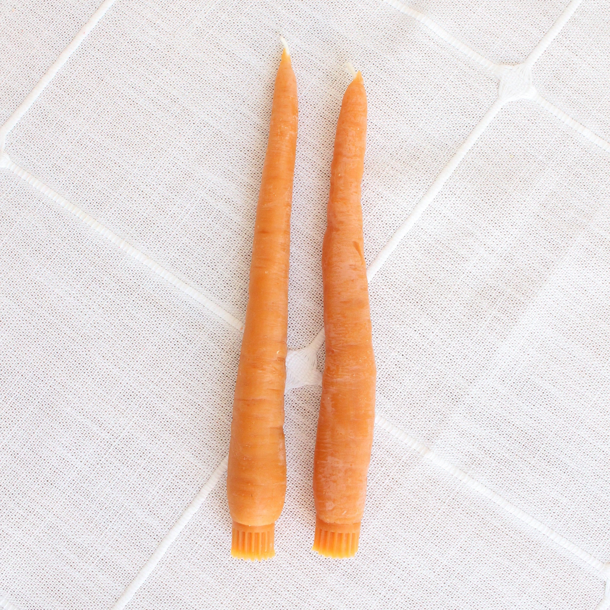 Two orange candles shaped like carrots on a textured white surface
