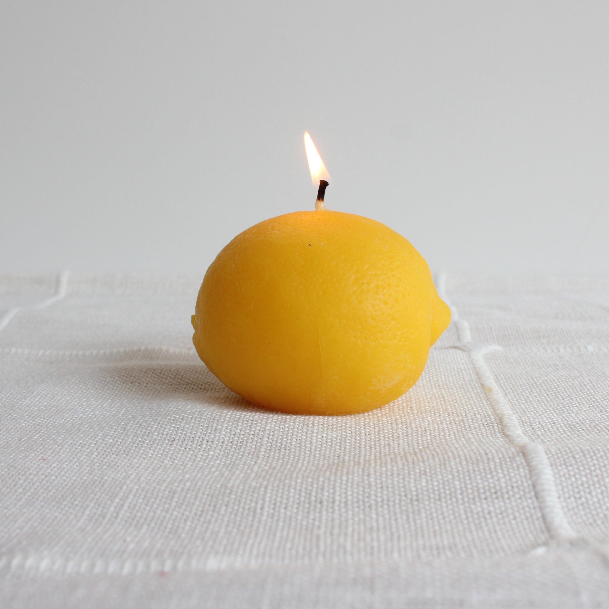 Yellow lemon-shaped candle with a lit wick on a white fabric background