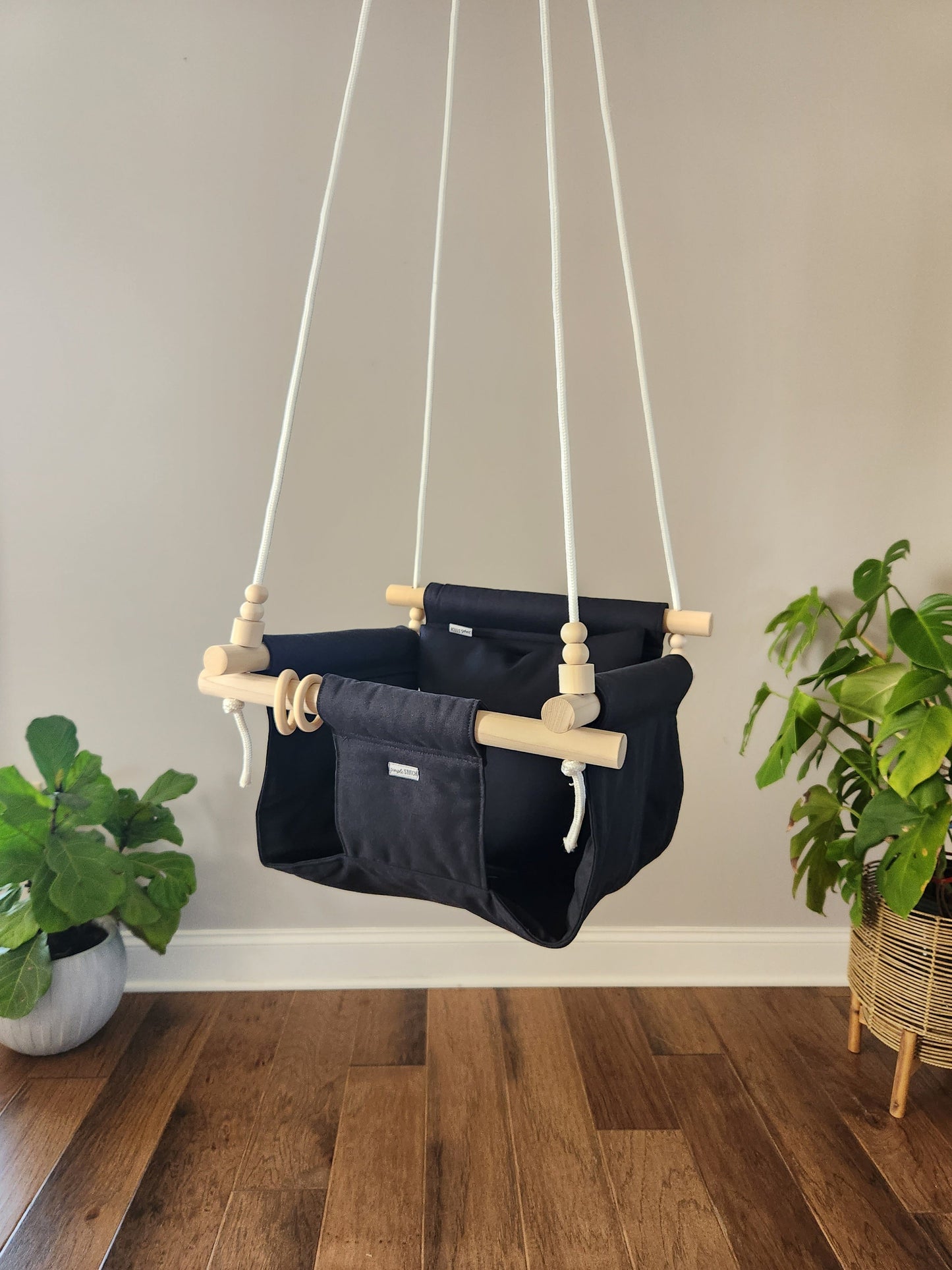 Navy baby swing with wooden handles hanging against a beige wall with two plants on either side.