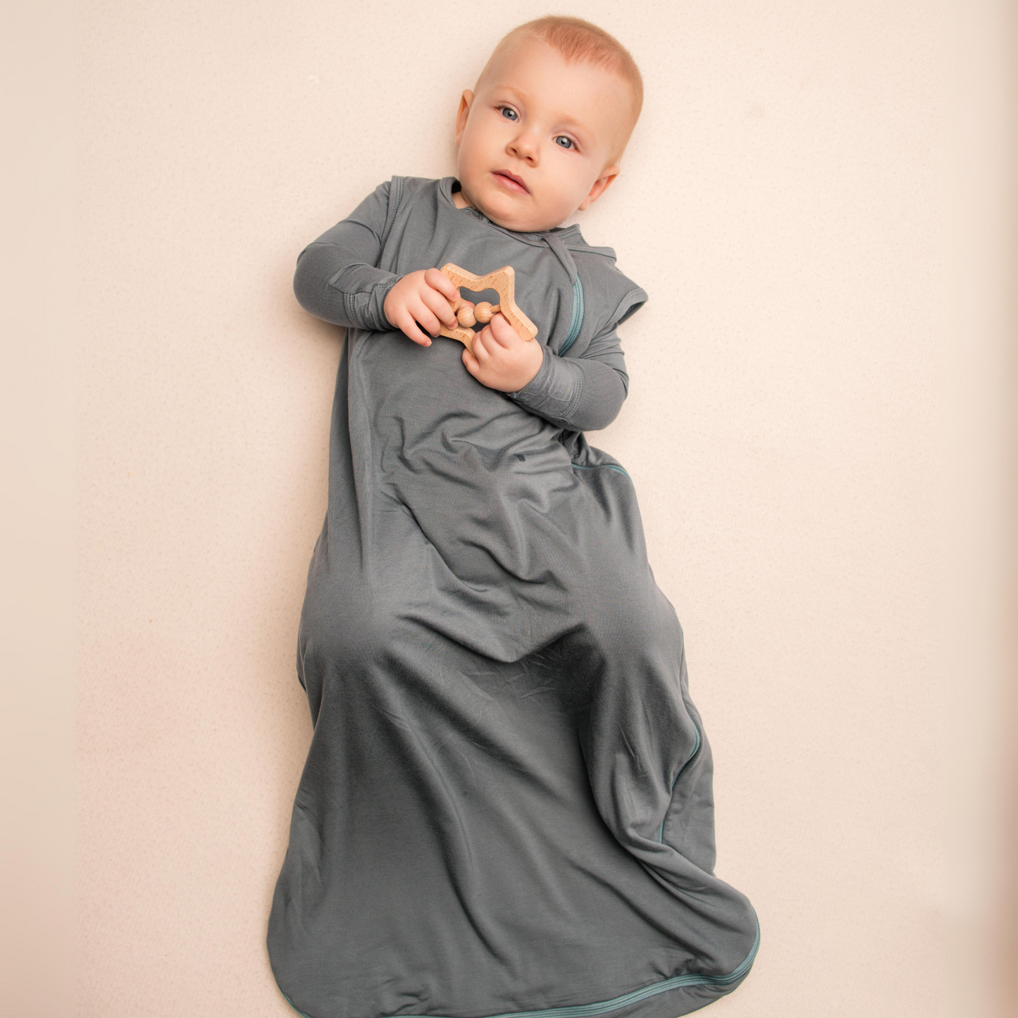 Baby in a gray swaddle holding a wooden toy against a beige background