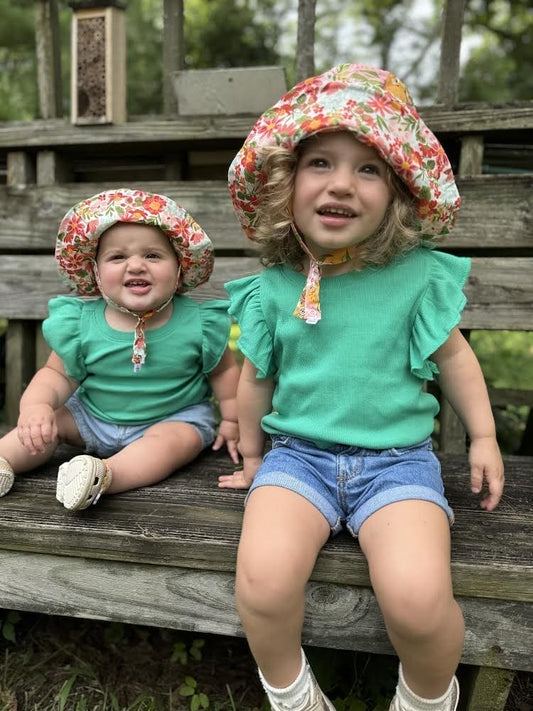 Two young girls wearing matching floral bonnets and green tops sitting on a wooden bench.