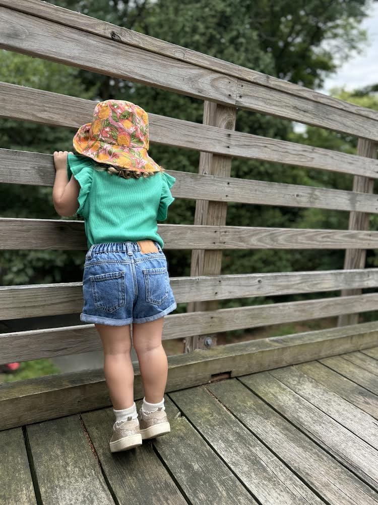Child wearing a floral hat, green shirt, and blue shorts standing on a wooden deck.