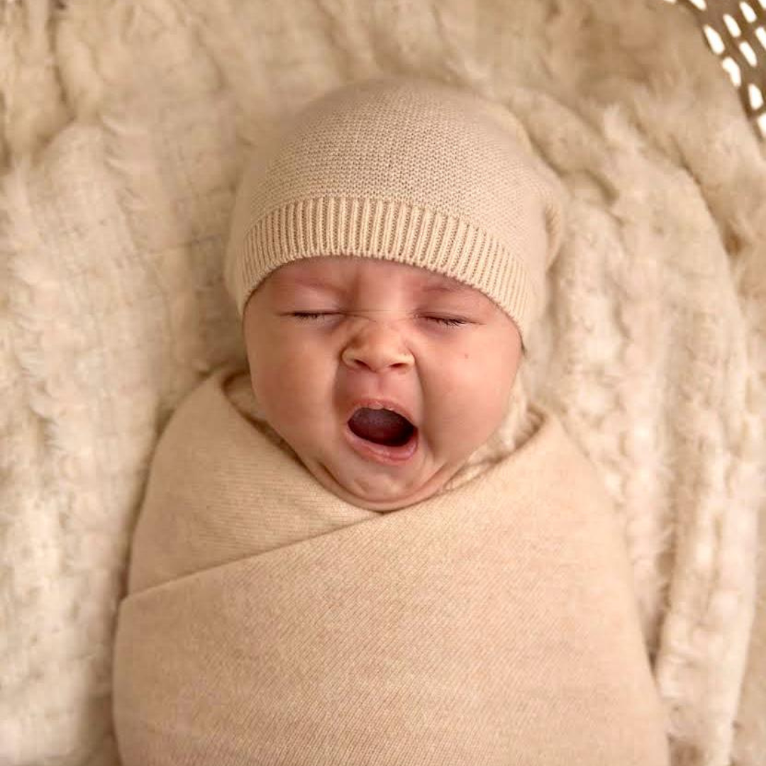 Newborn baby wrapped in beige fabric and wearing a matching hat, lying on a soft textured surface.