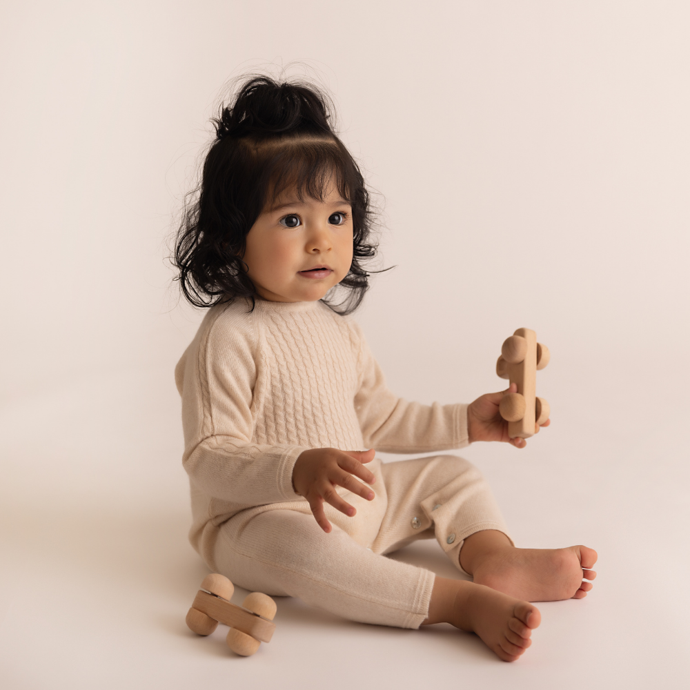 Baby in a beige outfit holding wooden toys on a plain background