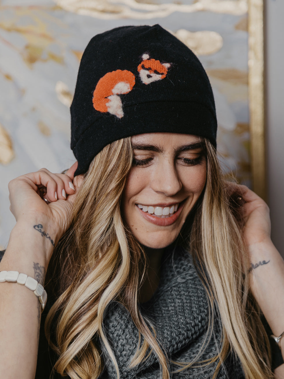Woman wearing a black beanie with fox designs, smiling outdoors.