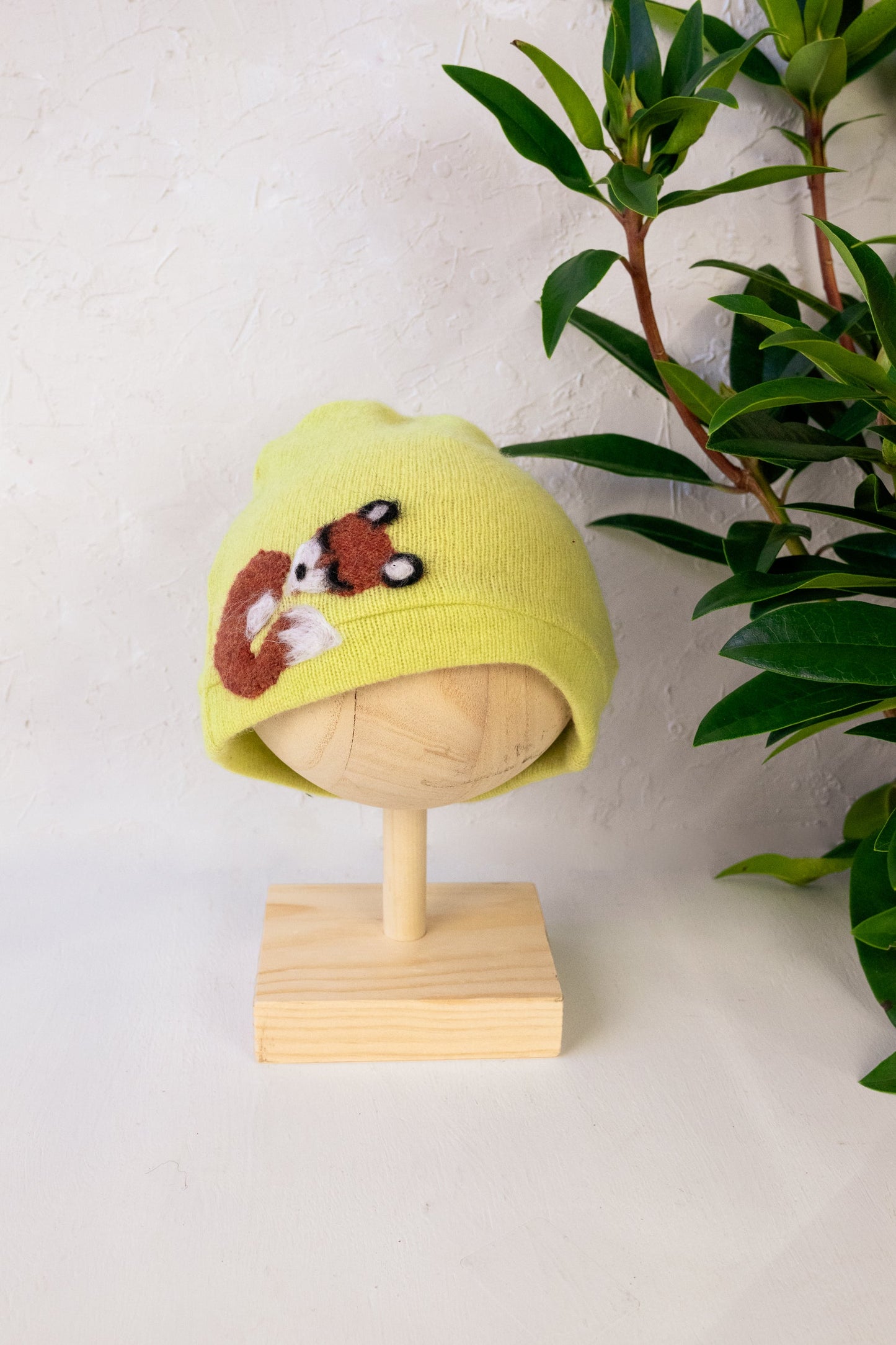 Yellow knitted hat with a bear design on a mannequin head against a white background with green leaves.