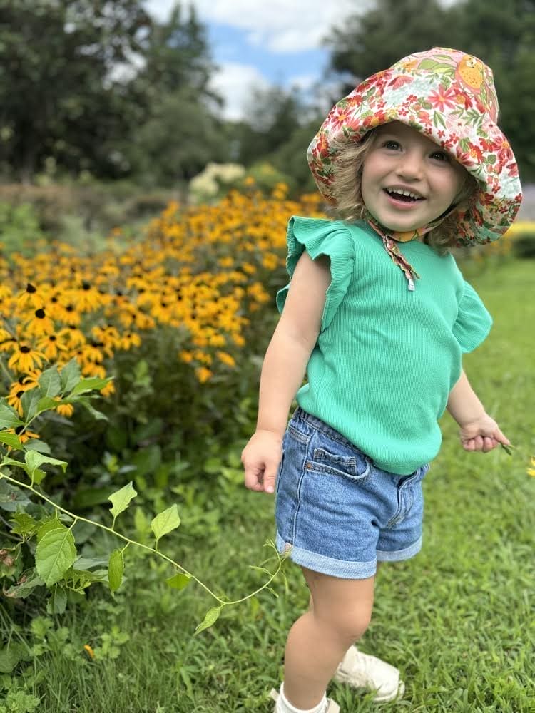 Child in a floral hat and green shirt standing in a garden with yellow flowers and greenery.