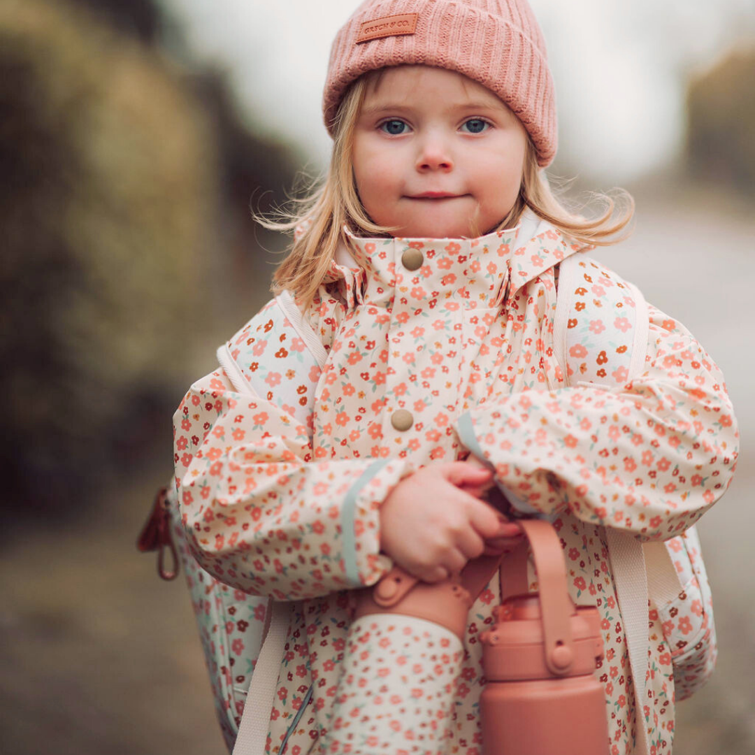 Child wearing a floral coat and pink hat holding a pink bag outdoors.