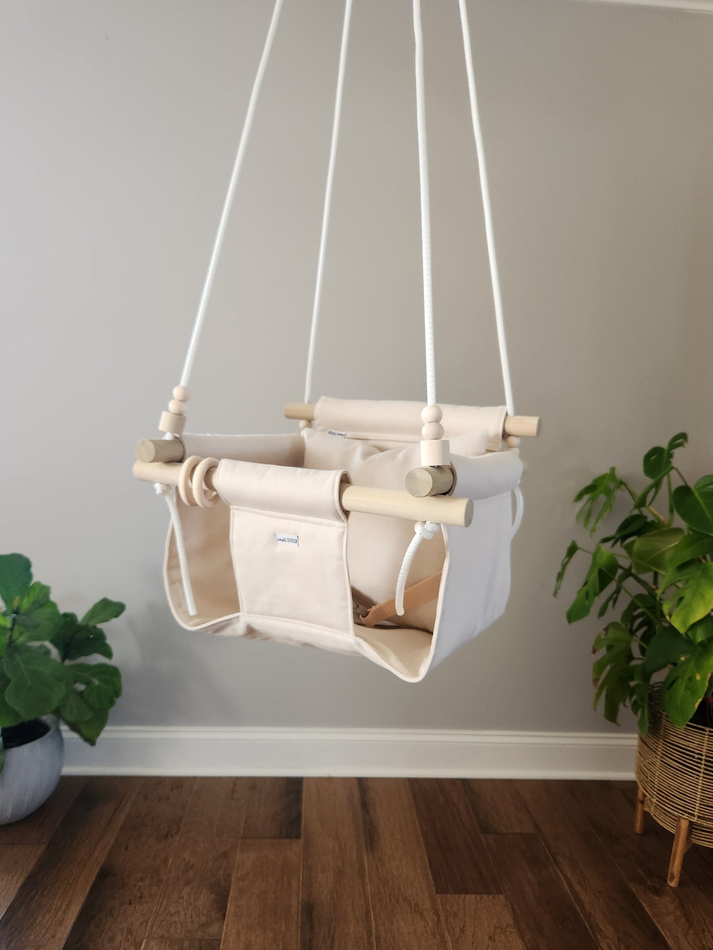 Children's swing hanging against a plain wall with plants on either side.
