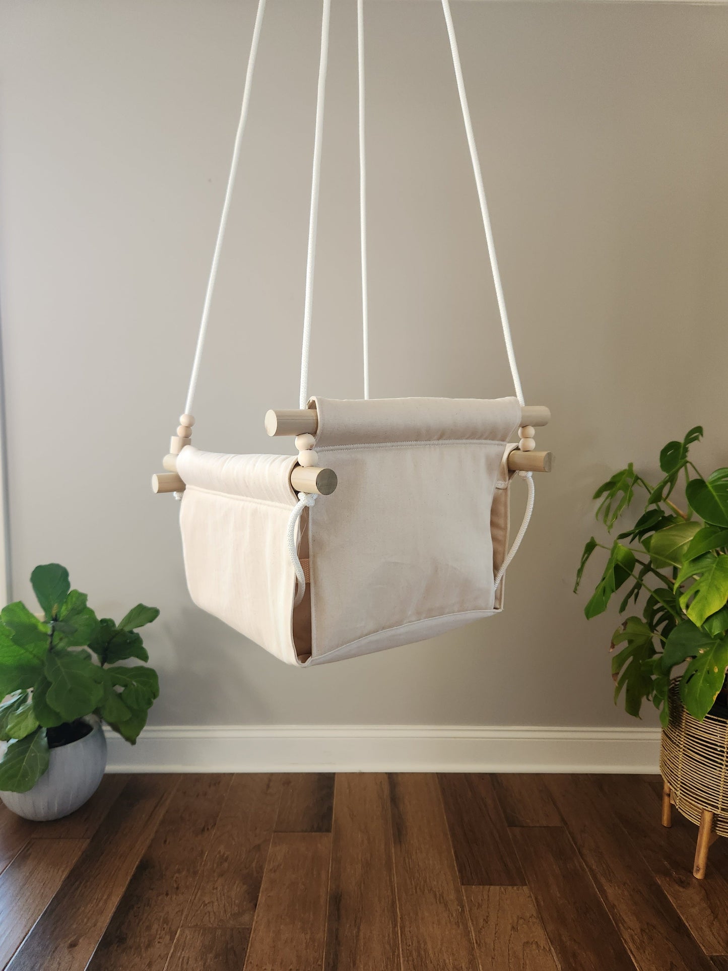 White baby swing hanging against a plain wall with plants on either side.