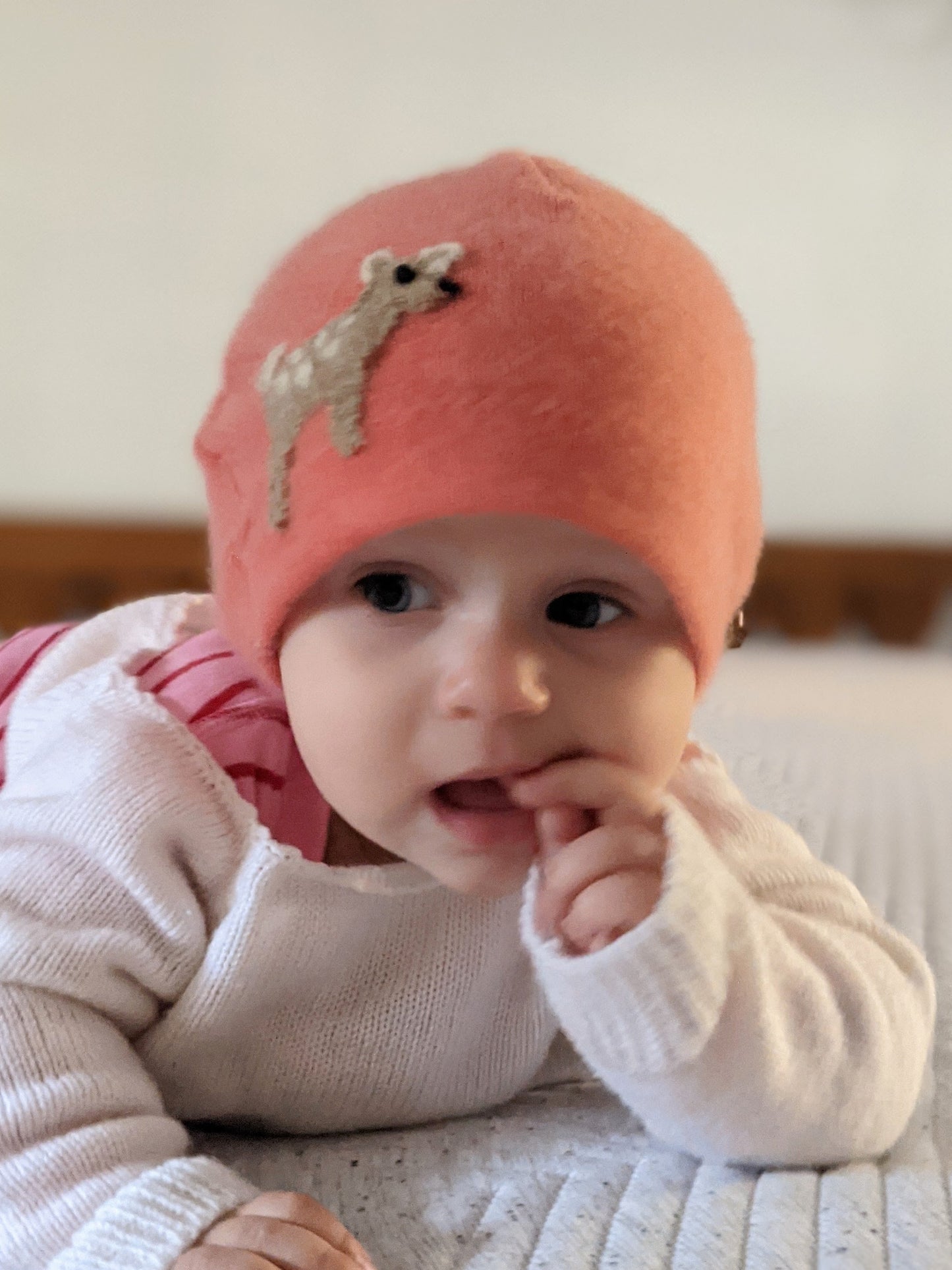 Baby wearing a pink knit hat with a deer design, sitting on a bed.