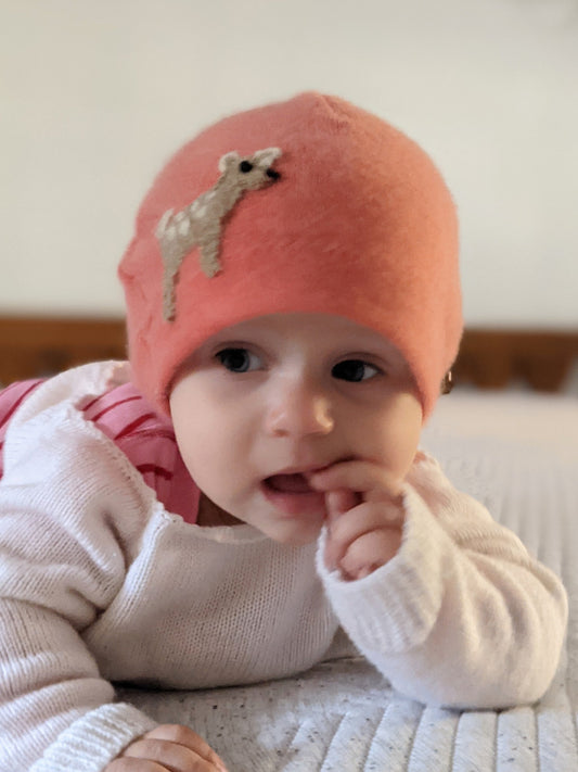 Baby wearing a pink knit hat with a deer design, sitting on a bed.